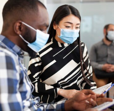 Diverse candidates wait in office lobby Closeup of asian woman reading application form of black man, both seated in office lobby waiting for interview meeting. Multiethnic male and female job candidates with face masks, sharing ideas.