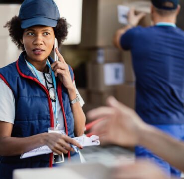 African American worker using phone while talking to her colleag Black female deliverer communicating with coworker while talking on mobile phone and organizing package delivery.